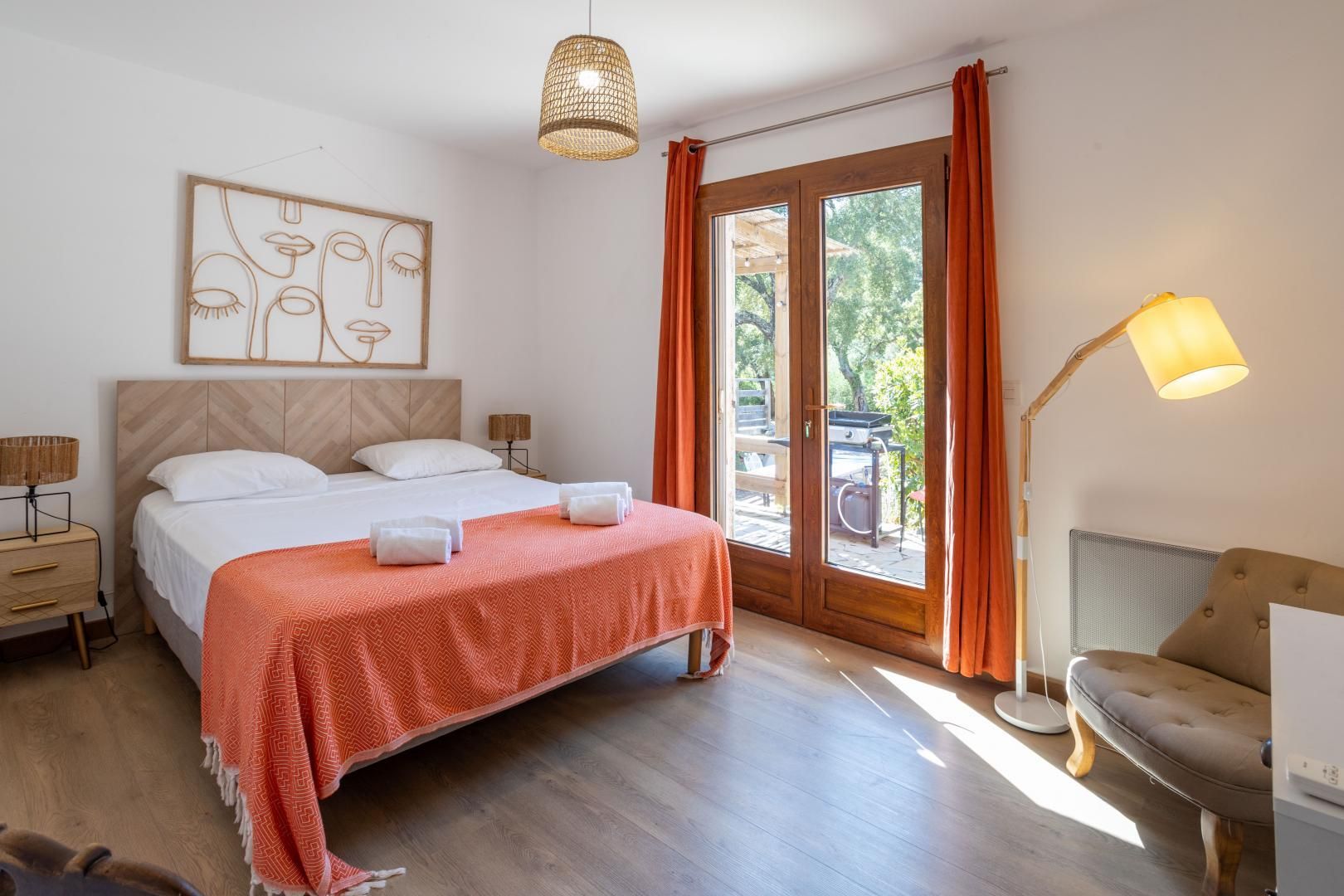 Ett hotellrum med en säng, ett sängbord, en lampa, en stol och utsikt över omgivningarna genom en glasdörr.