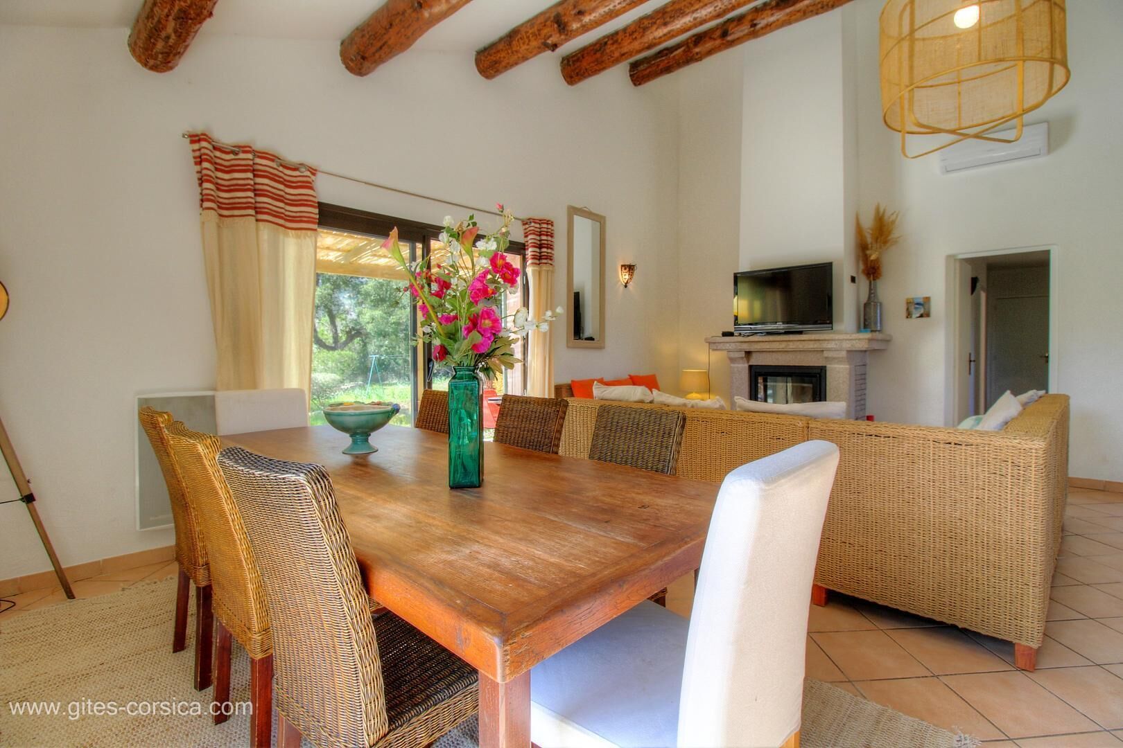 Un comedor con una mesa de madera, sillas de mimbre y un jarrón con flores.