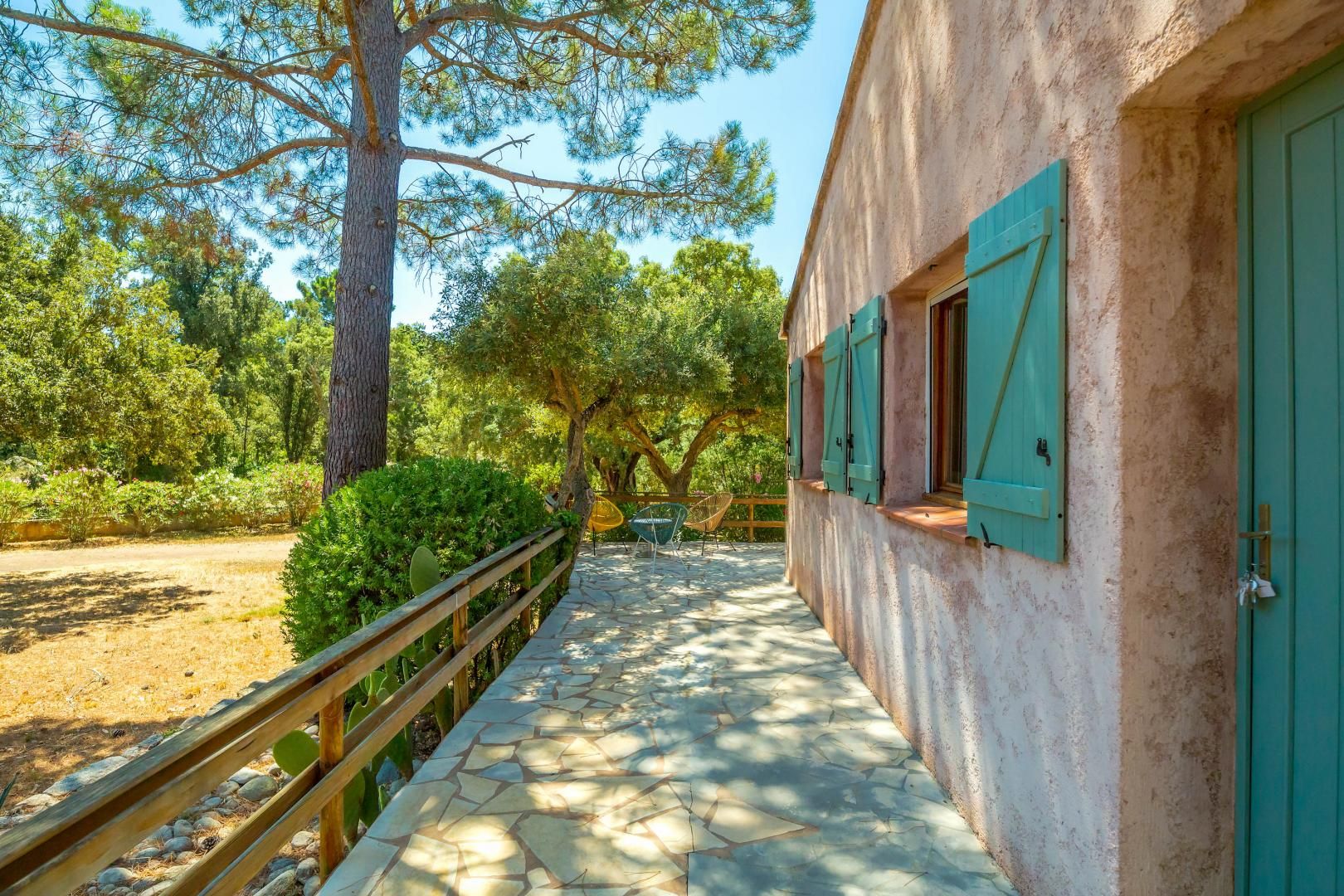 Un chemin de pierres mène à un bâtiment aux volets verts, entouré d’arbres et d’une clôture en bois.