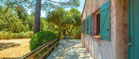 A stone pathway leading to a building with green shutters, surrounded by trees and a wooden fence.
