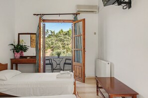 Een slaapkamer met een bed, nachtkastje, kapstok, spiegel, een potplant en een open deur naar een balkon met uitzicht op bomen.