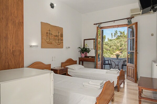 Slaapkamer met twee bedden, een bureau, een stoel, een potplant en een balkon met uitzicht op bomen.