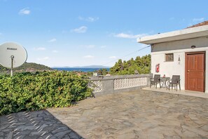Ein mit Steinplatten belegter Außenbereich mit einer Satellitenschüssel, einem Tisch mit Stühlen und Blick auf Meer und Hügel.