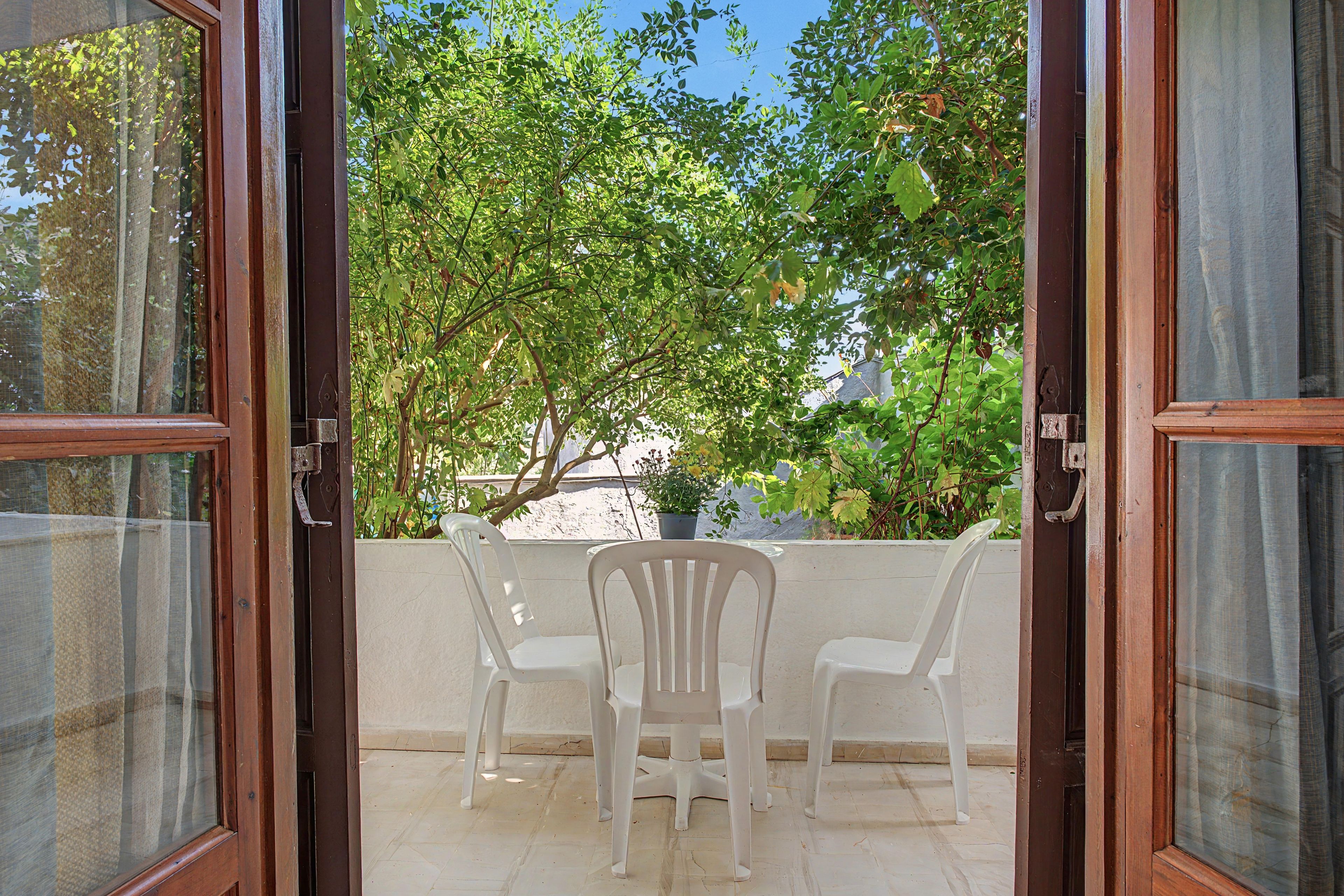 An open door leading to a patio with a white chair and a table, surrounded by greenery.