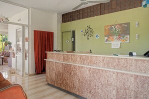 Ein Hotel-Empfangsbereich mit einer Marmor-Theke, einem Deckenventilator, einem Schild, das den Standort der Toilette anzeigt, und einer Wanddekoration mit Früchten und Blumen.