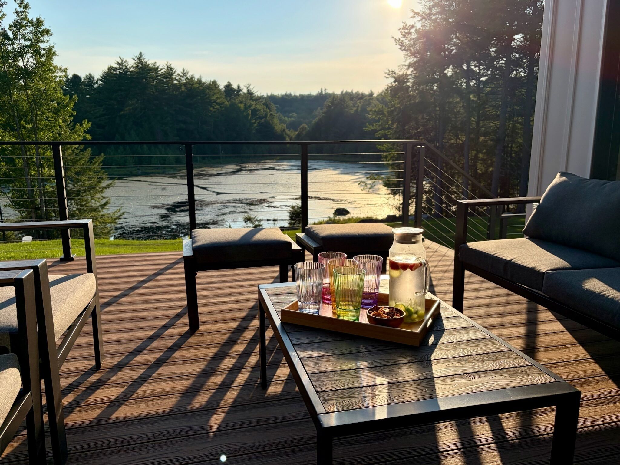 A deck with outdoor furniture, a tray with glasses and a pitcher, and a view of a lake.