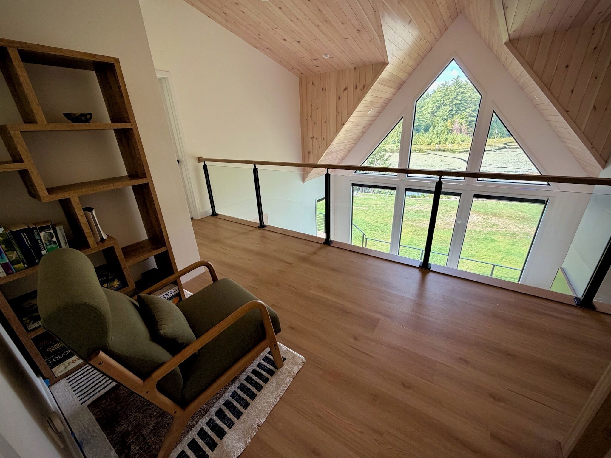 Un loft espacioso con techo alto, vigas de madera, un ventanal con vistas a un paisaje verde, una silla de madera y una estantería.