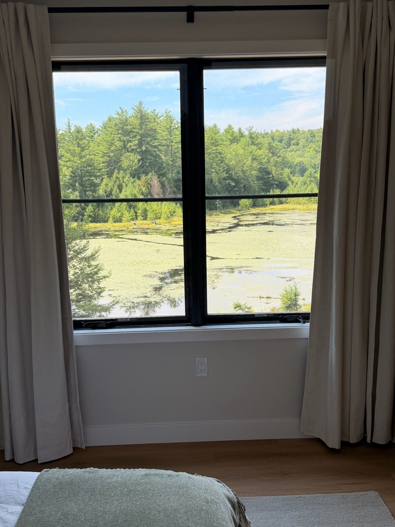 Una habitación con un ventanal, cortinas, una cama y vistas a un bosque y un campo abierto.