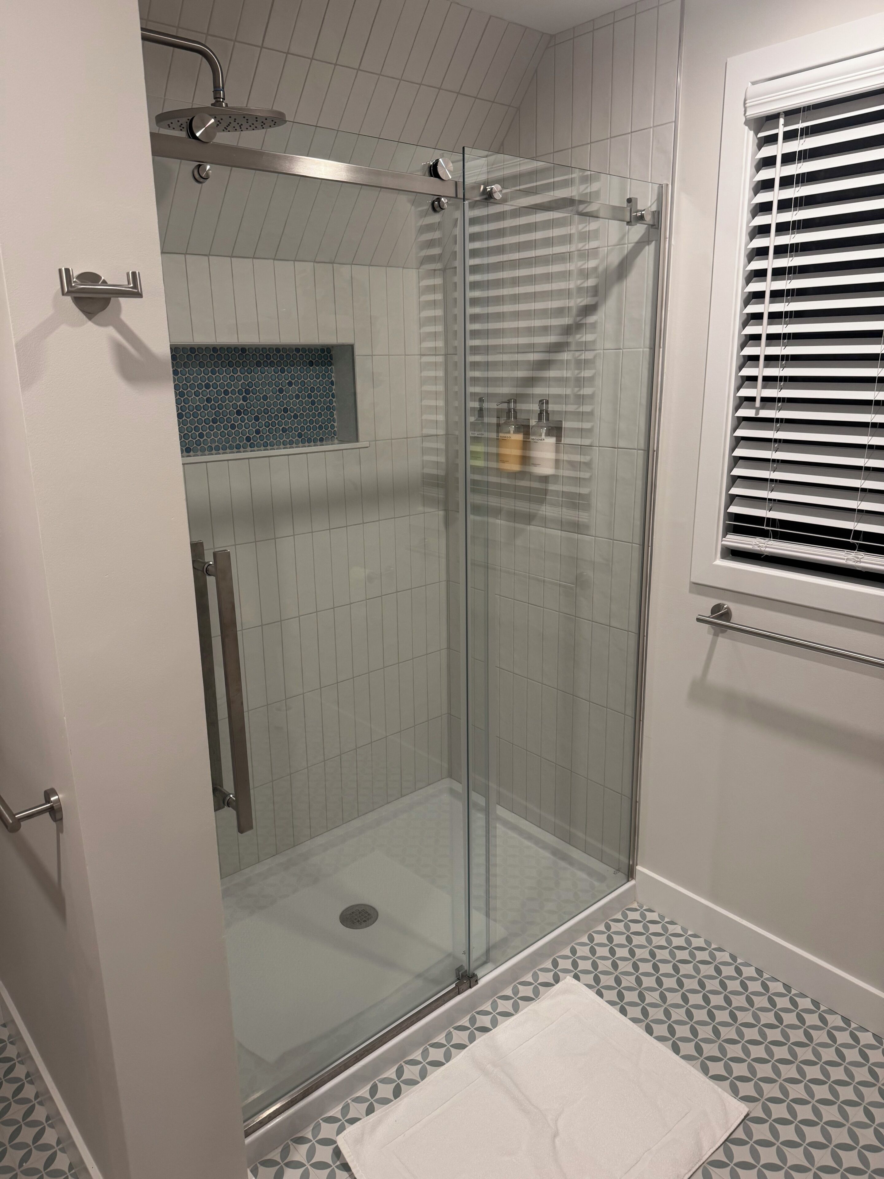 A glass-enclosed shower stall with a handheld showerhead, a towel on the floor, and a window with blinds.