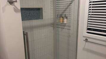 A glass-enclosed shower stall with a handheld showerhead, a towel on the floor, and a window with blinds.