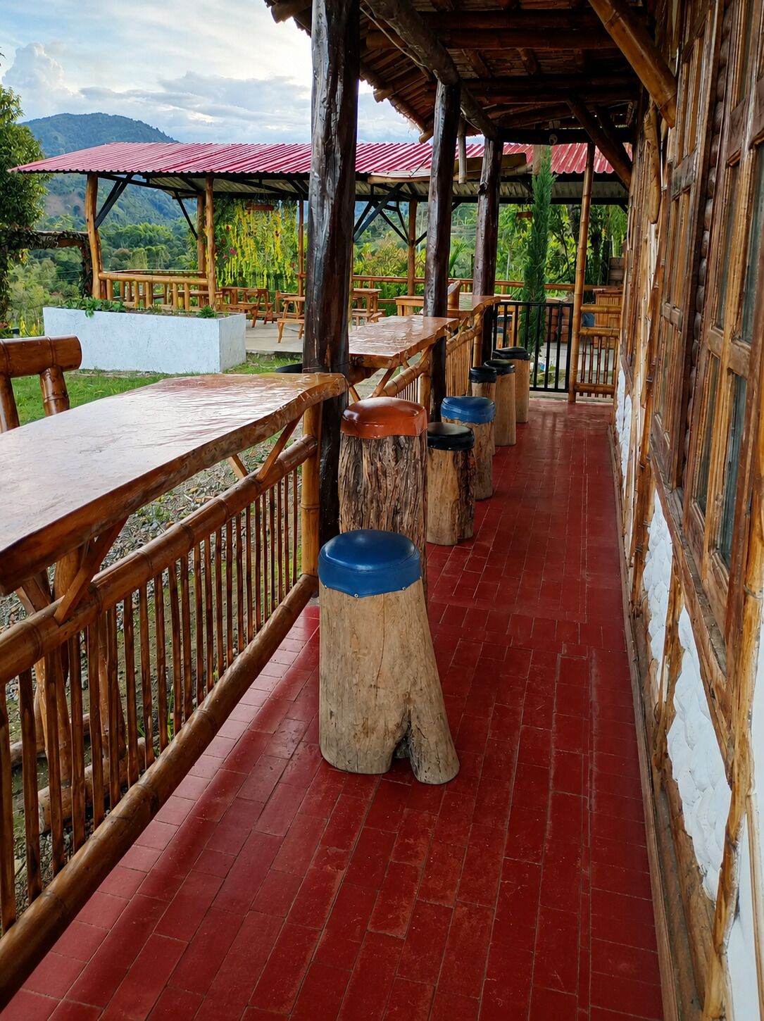 An outdoor covered patio area with wooden tables and benches, surrounded by a bamboo fence, and a view of greenery and hills in the background.