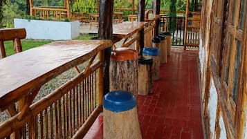 An outdoor covered patio area with wooden tables and benches, surrounded by a bamboo fence, and a view of greenery and hills in the background.