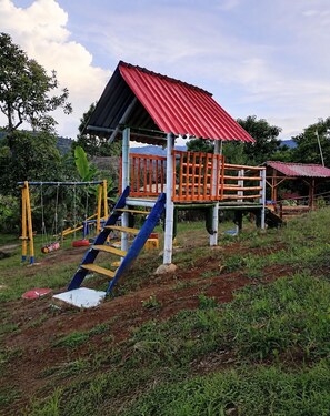 Un parque infantil con una casita de techo rojo, un tobogán azul y estructuras de escalada amarillas y anaranjadas.