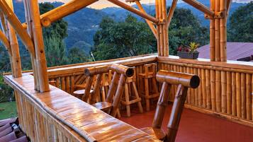 Estrutura de bambu com deck de madeira e área de estar, com vista para uma paisagem montanhosa.