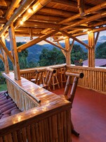 Estructura de bambú con terraza de madera y zona de estar, con vistas a un paisaje montañoso.