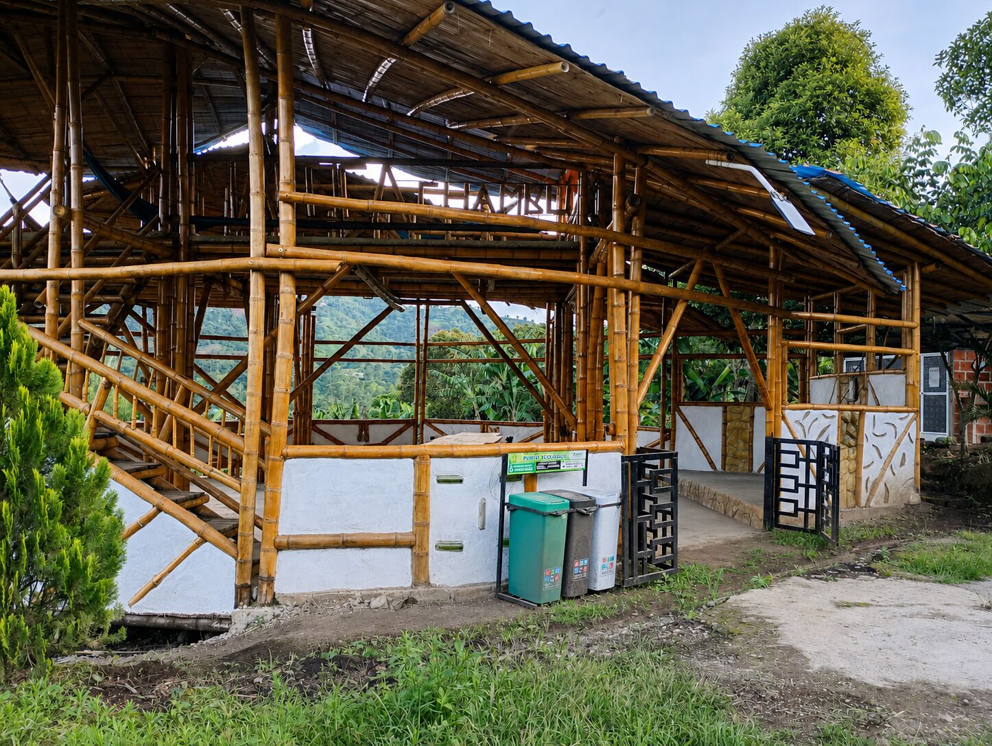 A bamboo-framed structure with a partially constructed roof, a small trash bin, and a gate.