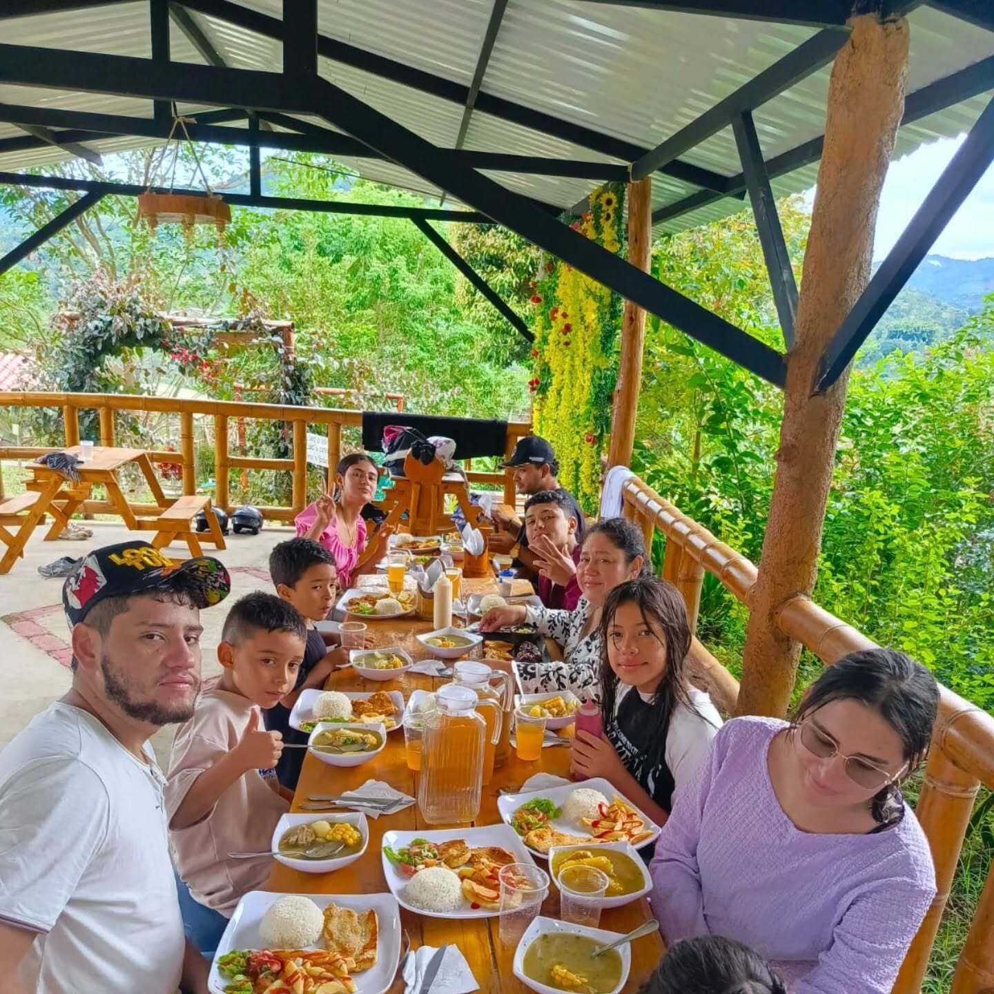 A group of people dining at an outdoor wooden table with various dishes and drinks, surrounded by lush greenery and a metal roof structure.