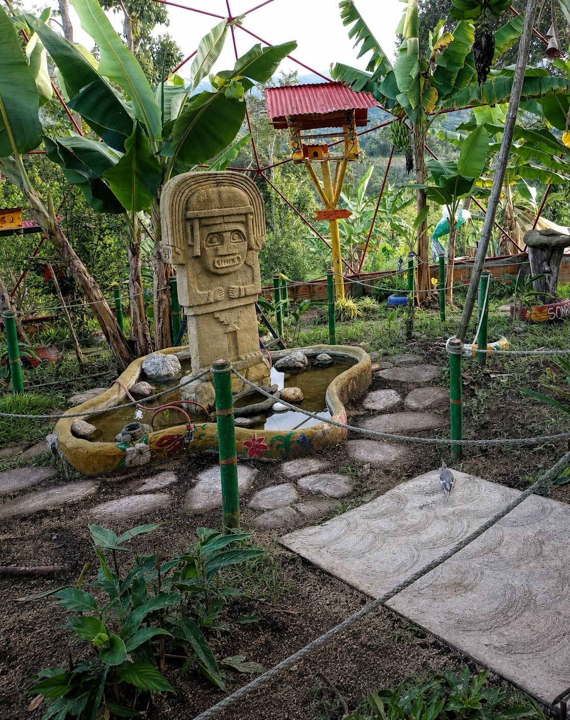 Una estatua de piedra con rostro, rodeada por un estanque con tortugas, ubicada en un jardín con palmeras y una pequeña estructura de techo rojo.