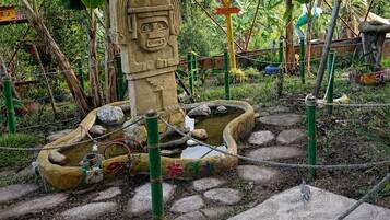 Una estatua de piedra con rostro, rodeada por un estanque con tortugas, ubicada en un jardín con palmeras y una pequeña estructura de techo rojo.