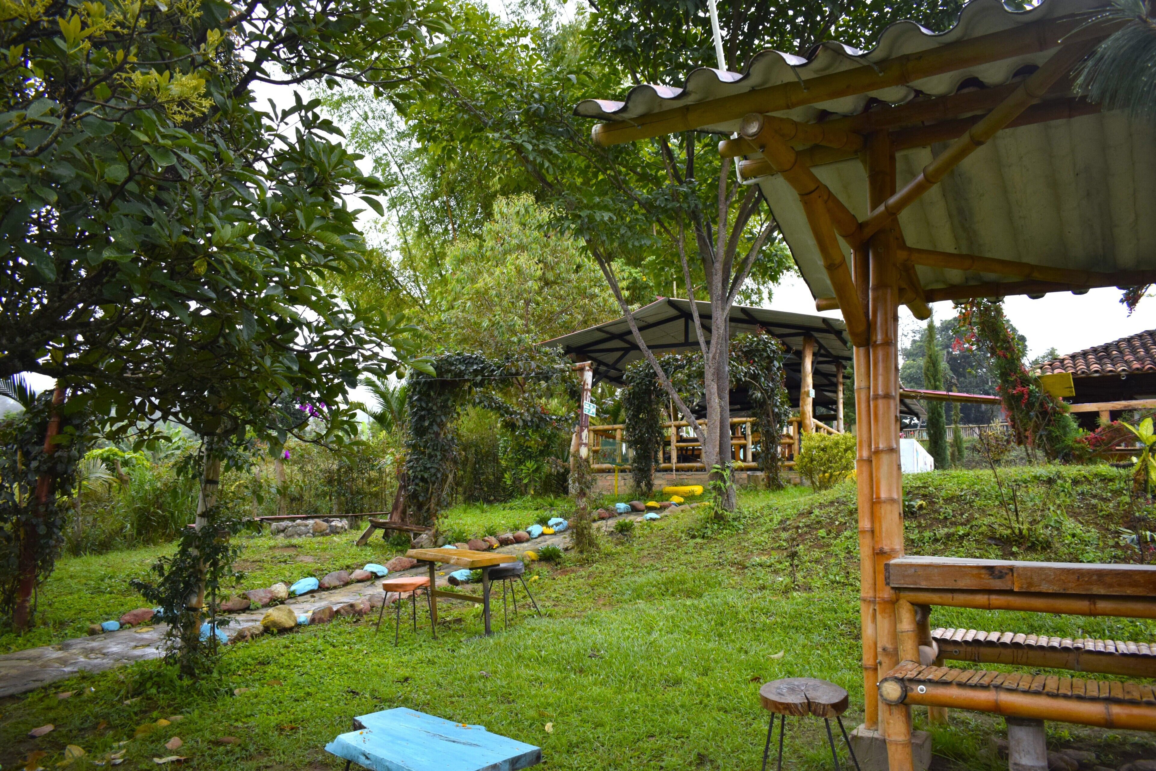 Un espacio de estar al aire libre cubierto, con estructura de madera, rodeado de abundante follaje y un sendero de piedra.
