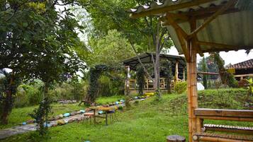 Un espacio de estar al aire libre cubierto, con estructura de madera, rodeado de abundante follaje y un sendero de piedra.