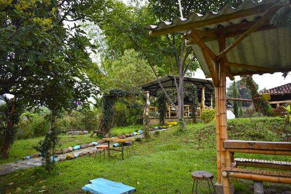 Un espace extérieur aménagé avec des sièges, abrité par une structure en bois, entouré d’une végétation luxuriante et bordé d’un sentier en pierre.