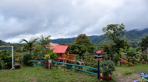 Un parque infantil con una estructura de techo rojo, rodeado de abundante vegetación y un paisaje montañoso.
