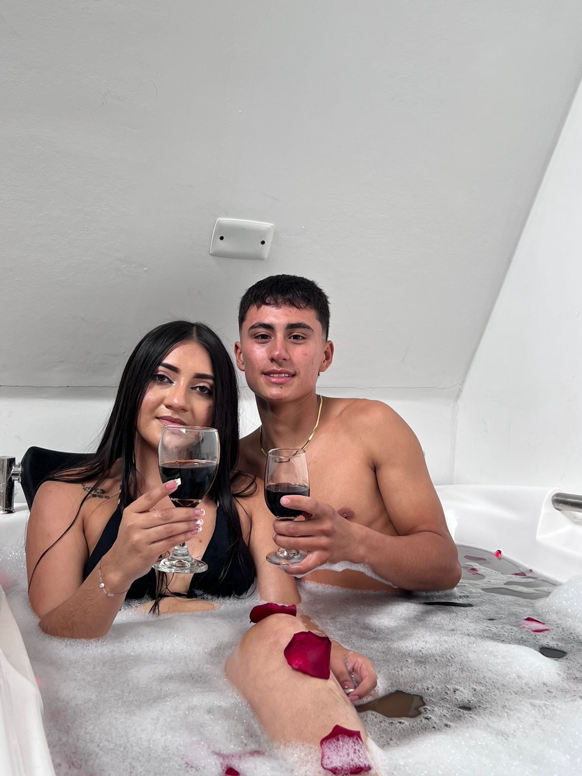 Two individuals in a bathtub with rose petals, holding glasses of red wine.