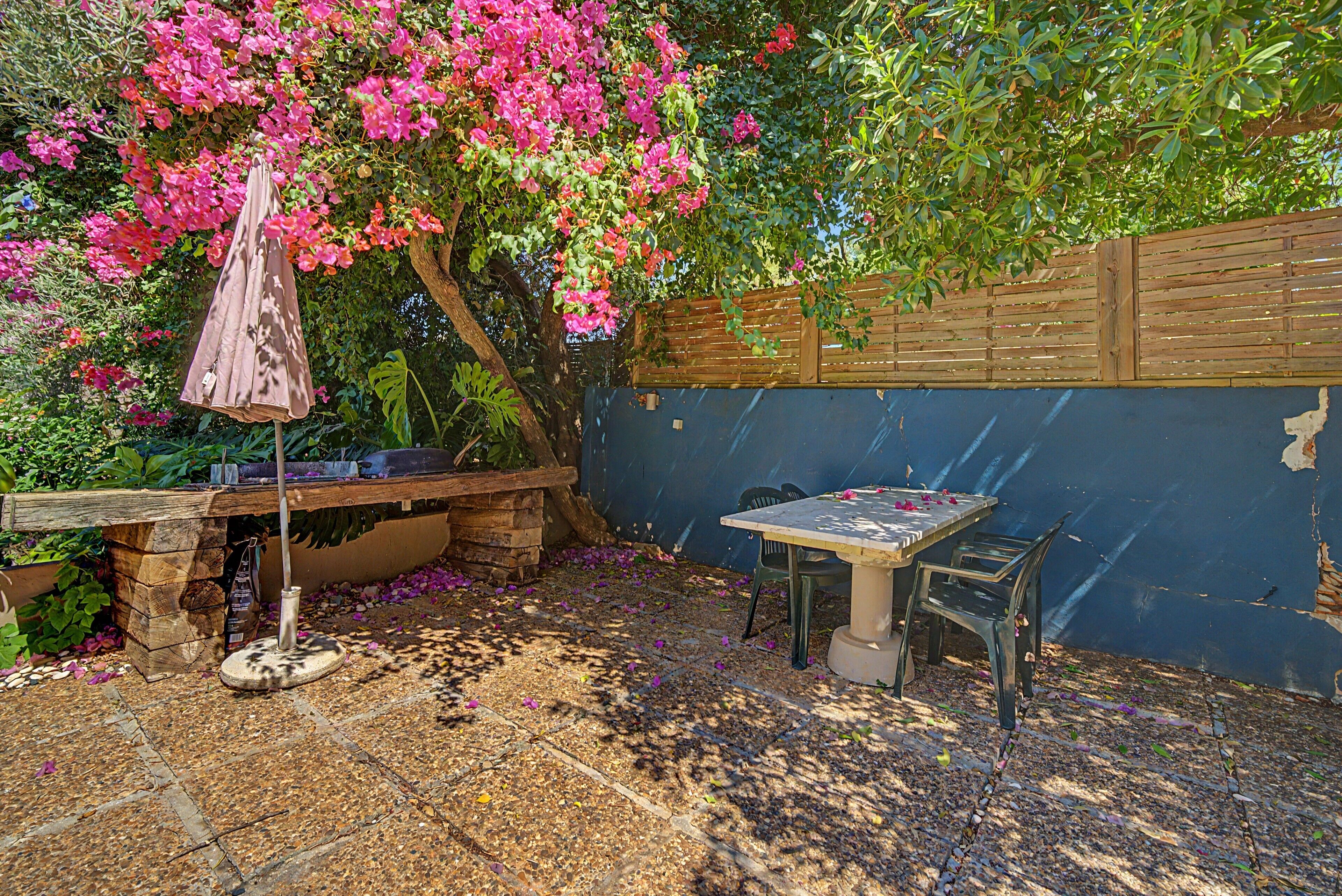 A patio area with a table and chairs, surrounded by a wooden fence and a garden with pink flowers.