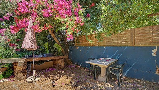Un patio con mesa y sillas, rodeado por una valla de madera y un jardín con flores rosadas.