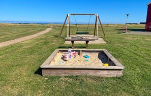 A playground with a sandbox, swings, and a red building in the background. - 2 bedroom gorgeous home in Hadsund (Hadsund)