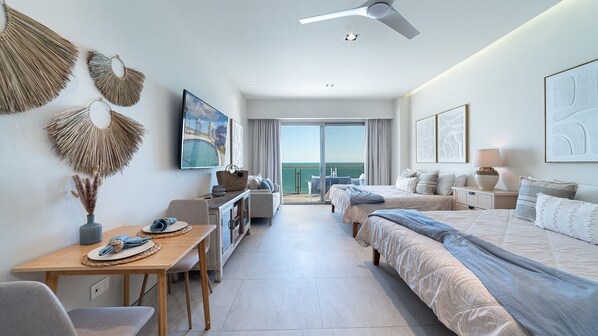 Ein Hotelzimmer mit Bett, Schreibtisch, Stuhl, Tisch, Fernseher und einem Balkon mit Meerblick.