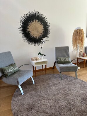 Ein modernes Wohnzimmer mit zwei grauen Sesseln, einem runden grauen Teppich, einem weißen Beistelltisch mit einer Pflanze und einem großen abstrakten Wandbild.