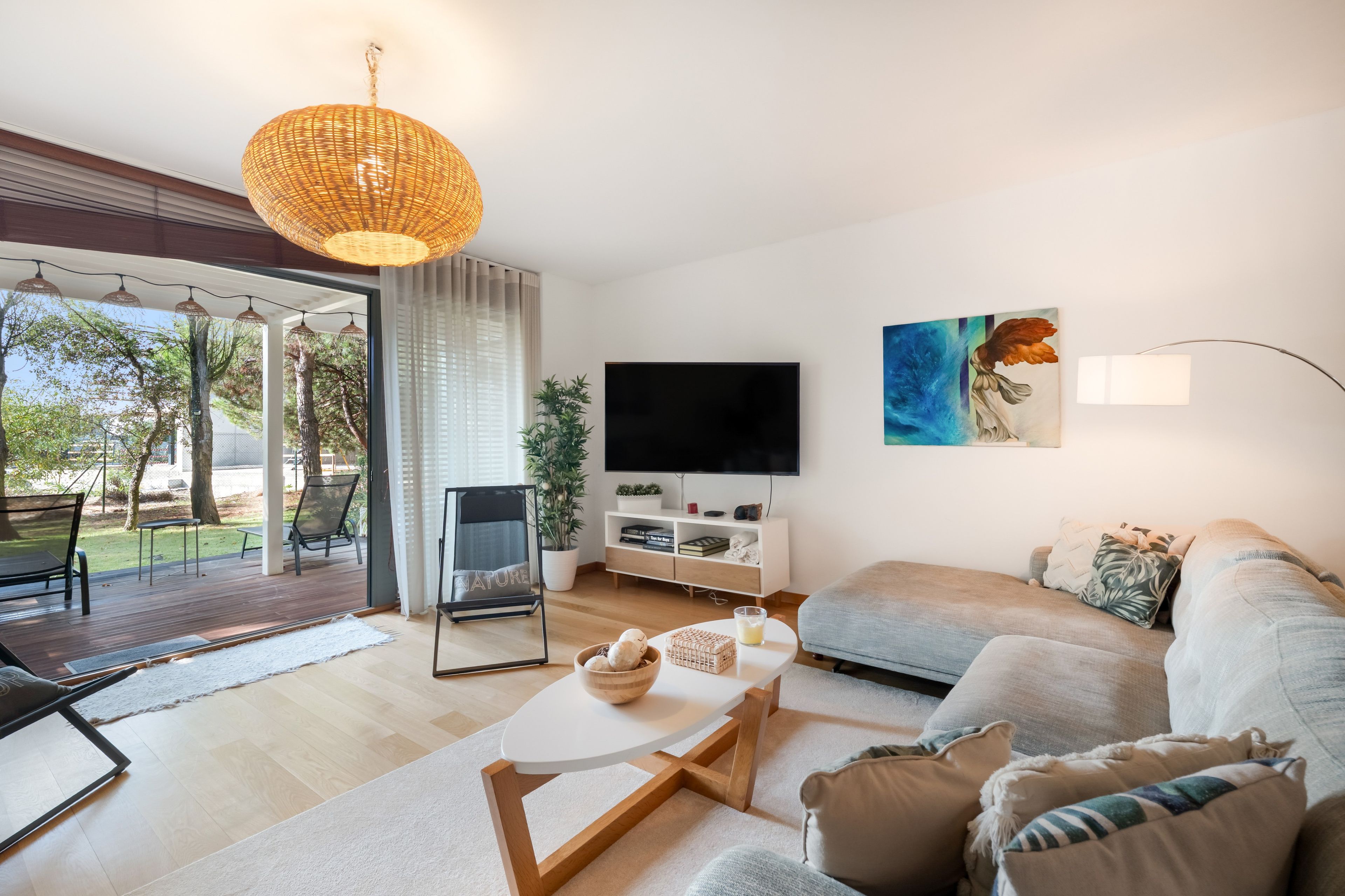 Una sala de estar moderna con sofá, mesa de centro y televisor. Hay un balcón con una silla y una mesa, y una ventana grande con cortinas.