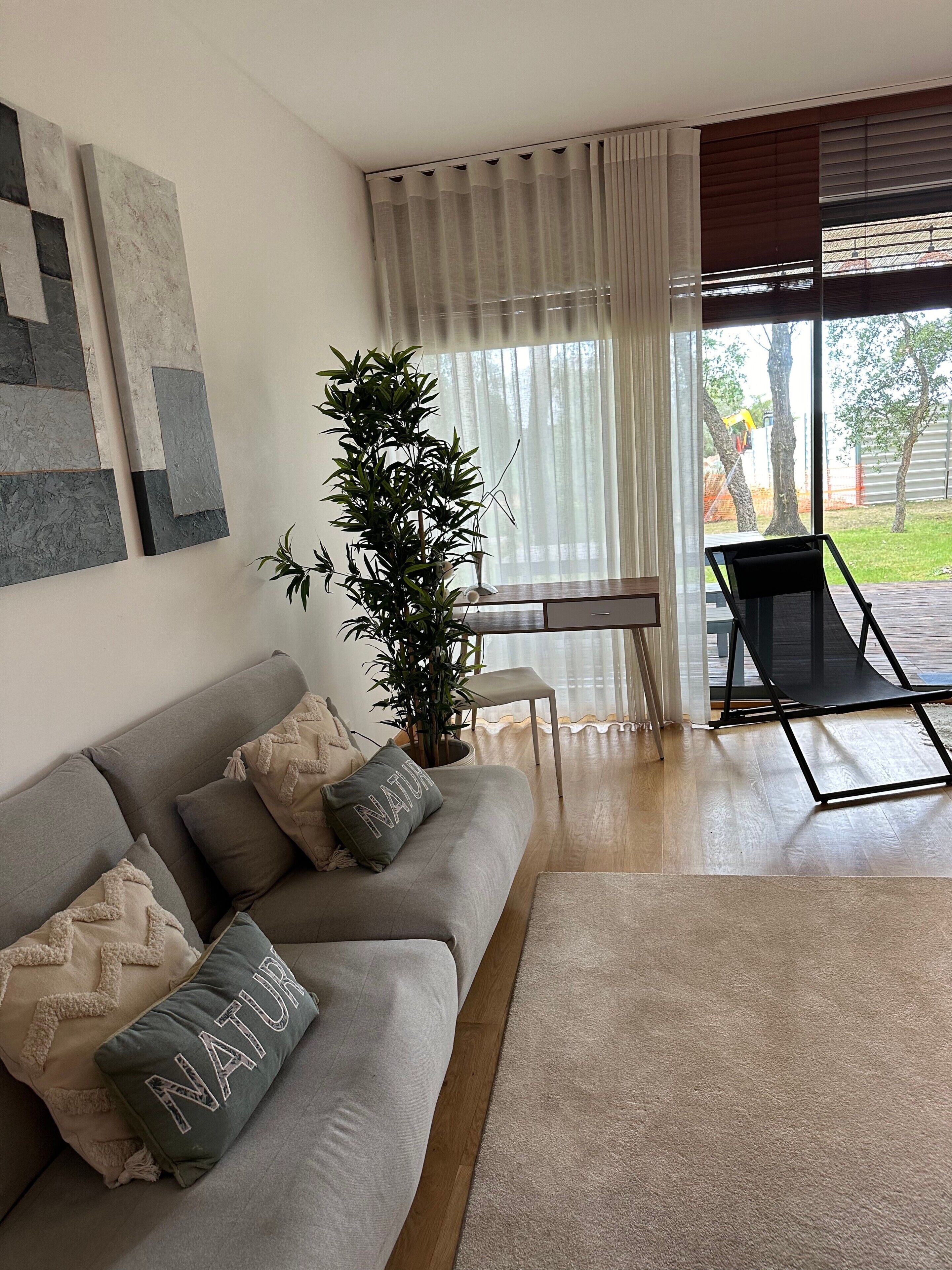 A modern living room with a grey sofa, a potted plant, and a view through large windows.