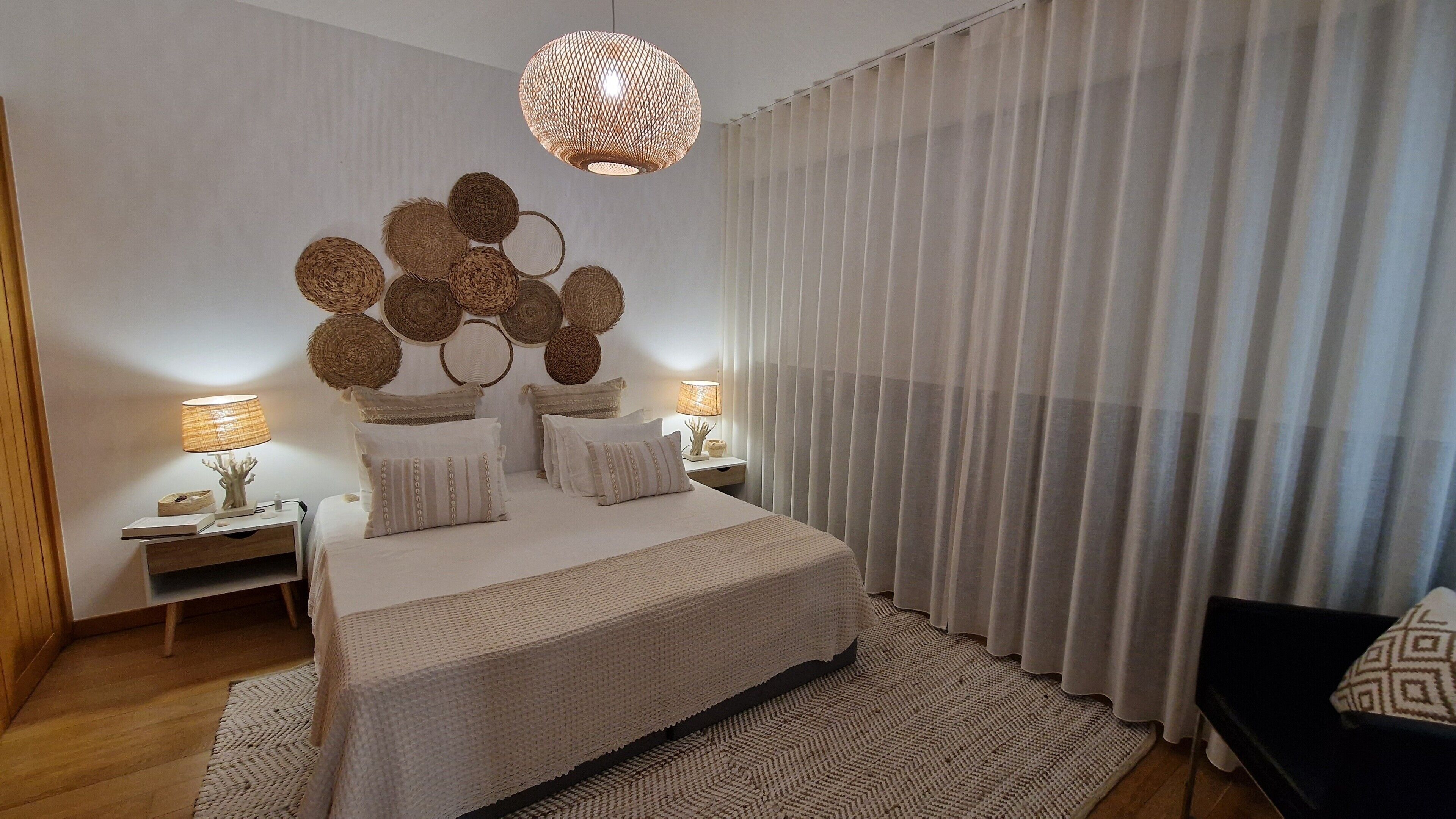 Een slaapkamer met een groot bed, twee nachtkastjes, een hanglamp en een raam met dunne gordijnen.