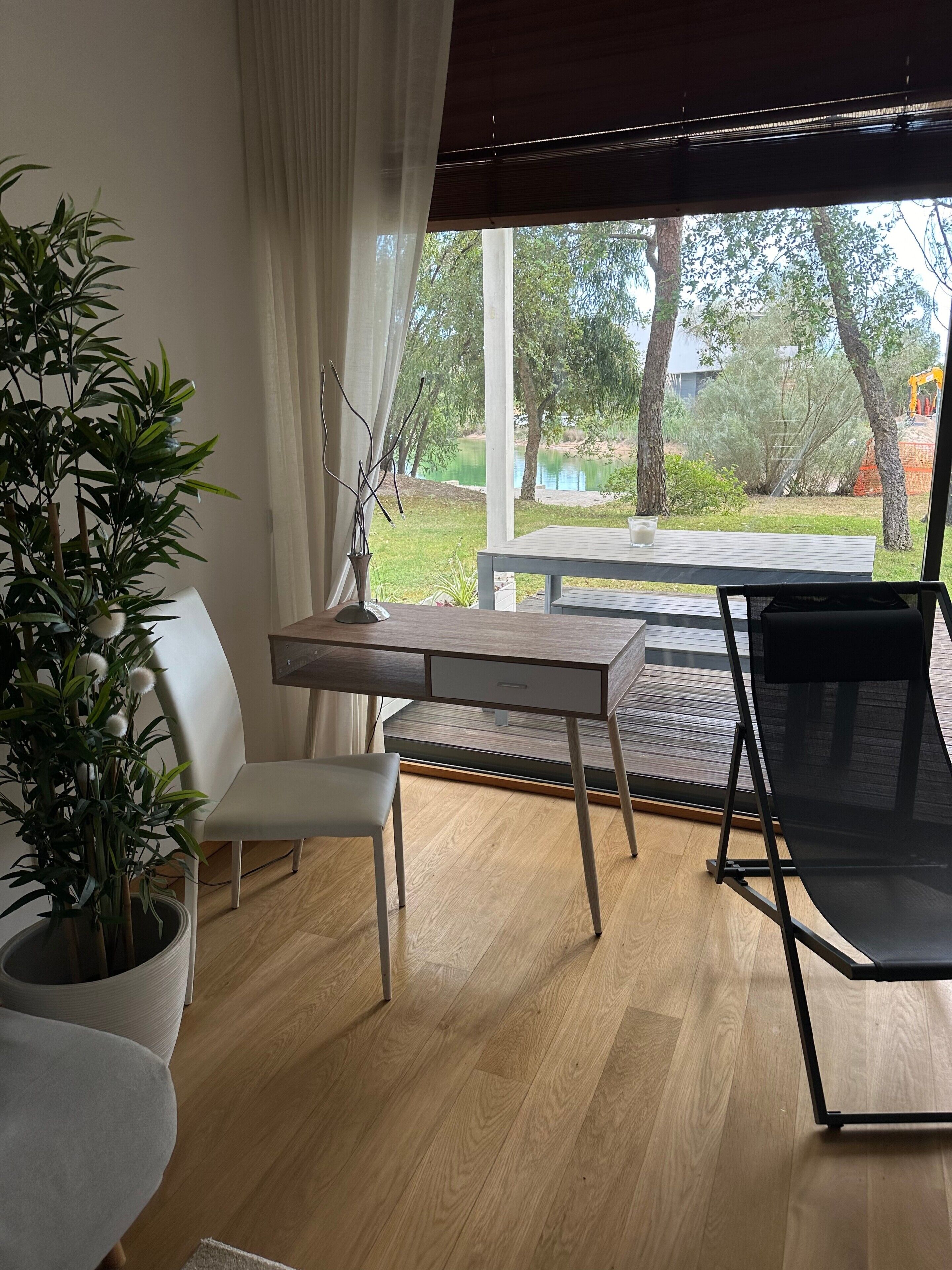Een kamer met een houten bureau, een witte stoel, een zwarte stoel, een potplant en uitzicht op een tuin en een meer.