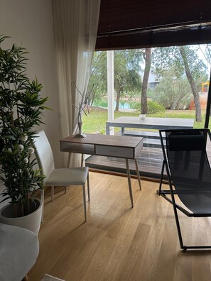 A room with a wooden desk, a white chair, a black chair, a potted plant, and a view of a garden and lake.