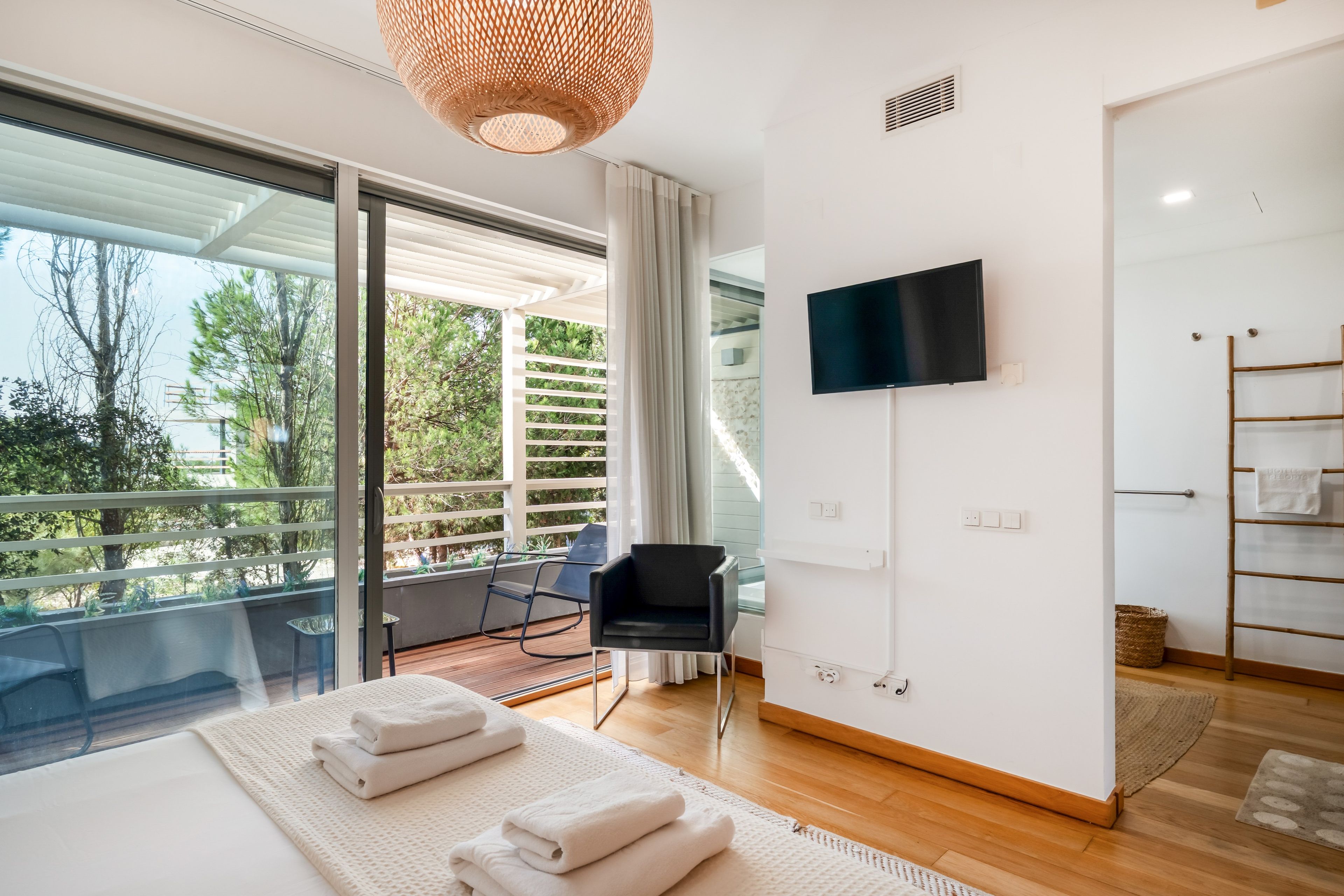 Un dormitorio moderno con un ventanal grande, un televisor de pantalla plana, una cama con toallas ordenadas y un balcón con una silla.