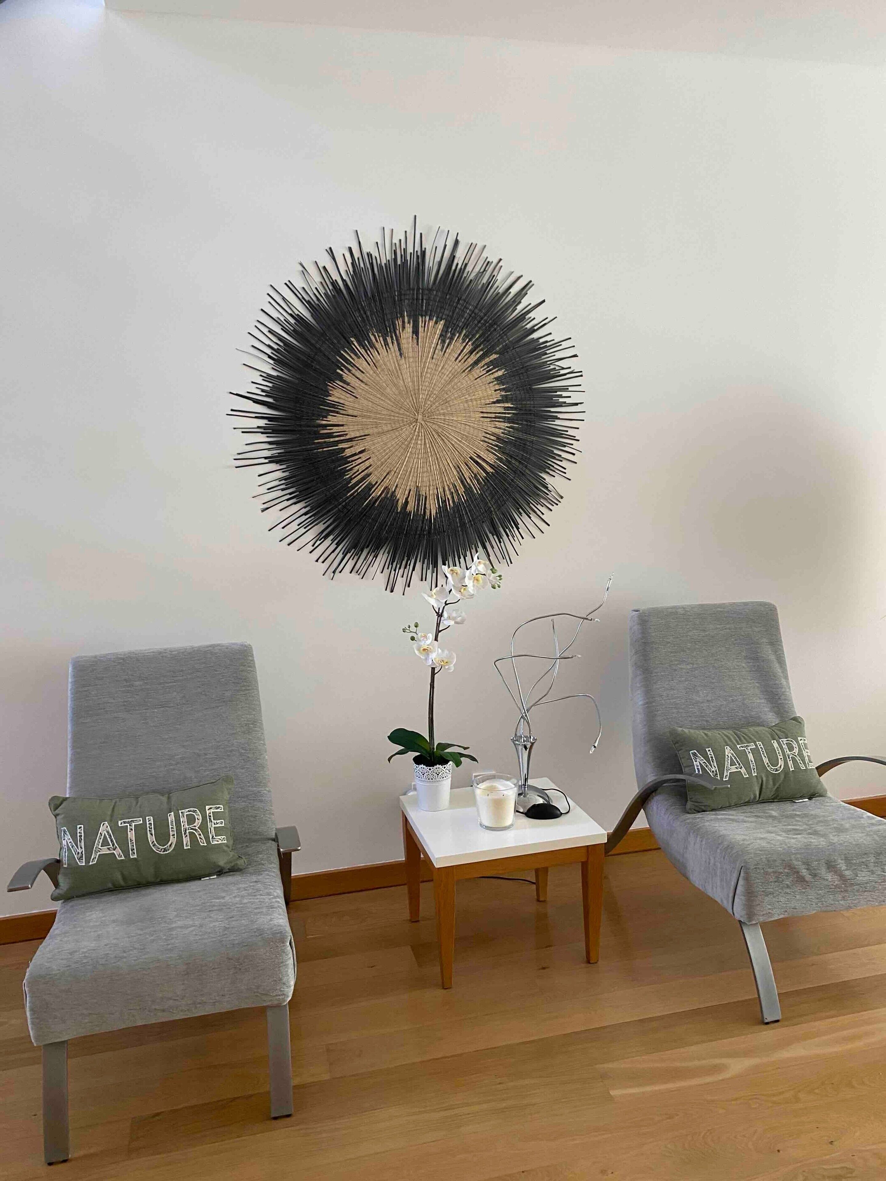 Twee grijze fauteuils met kussens met de tekst 'NATURE', een witte tafel met een kaars en orchidee, en een zwart-beige wanddecoratie.