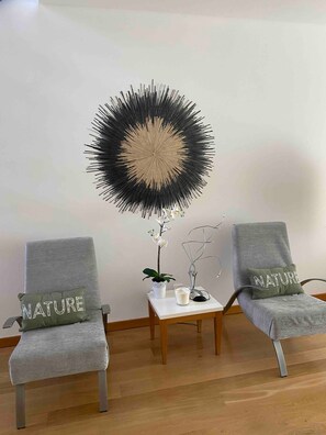 Two grey armchairs with 'NATURE' pillows, a white table with a candle and orchid, and a black and beige wall art piece.