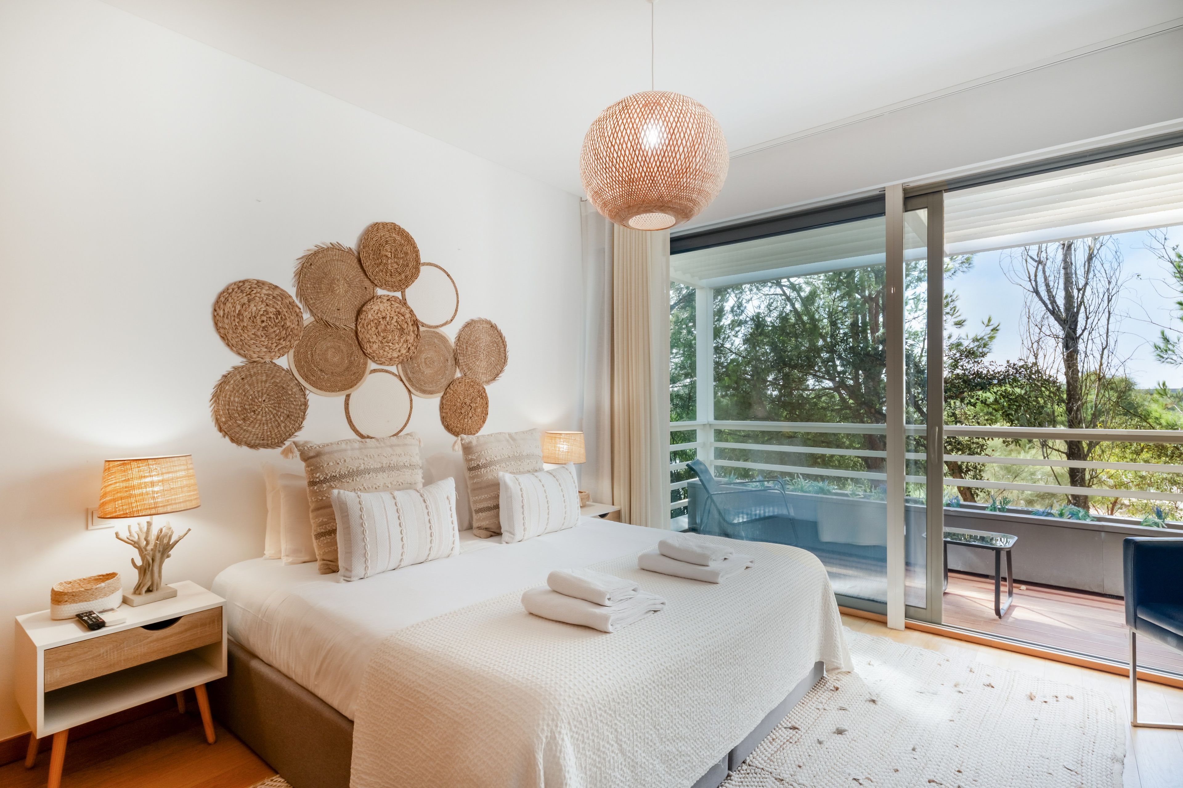 A modern bedroom with a large bed, a decorative wall hanging, a bedside table with a lamp, and a view of the outdoors through a sliding glass door.