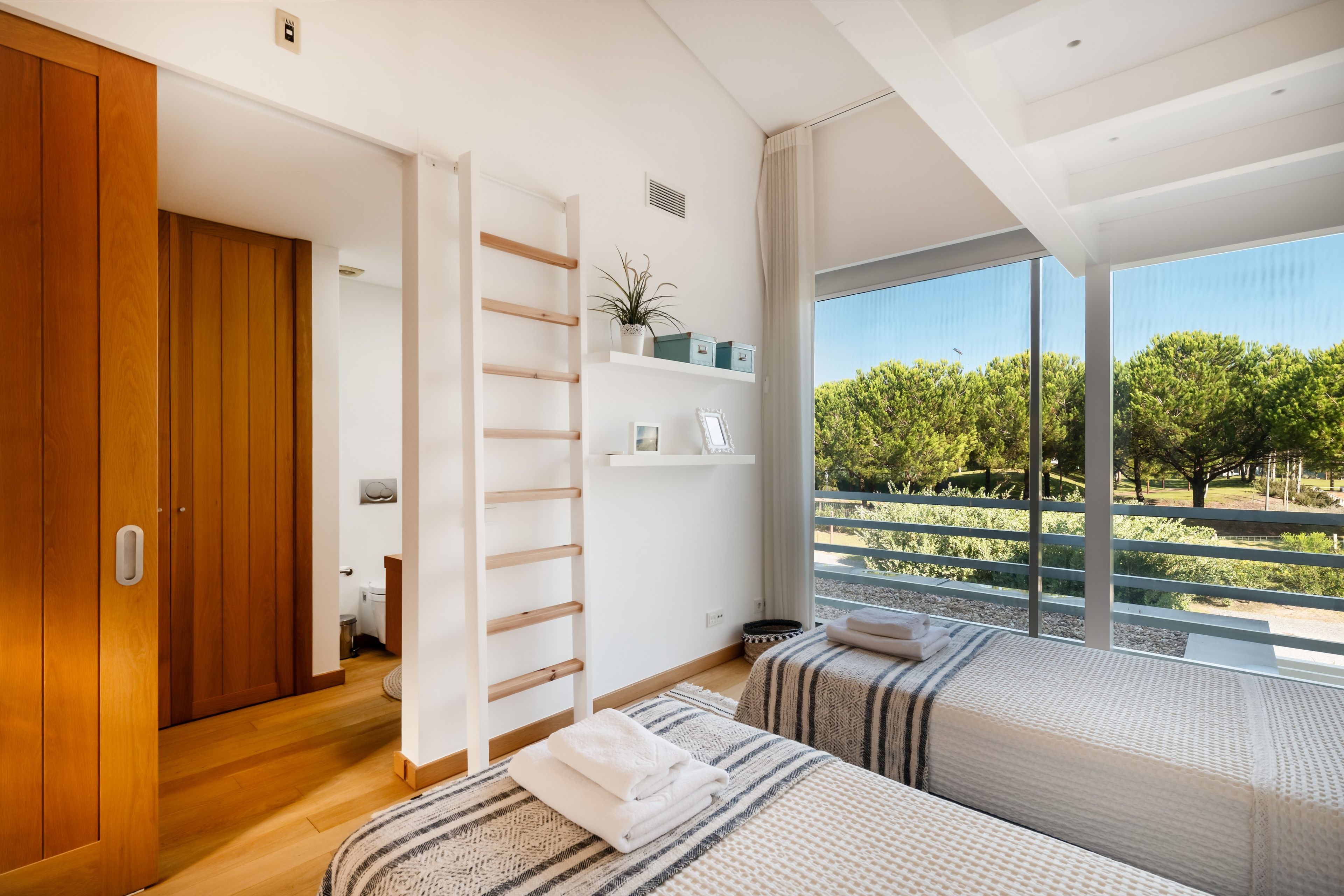 A modern bedroom with a wooden wardrobe, a built-in shelf with decorative items, a large window with a view of trees, and a bed with a striped blanket.