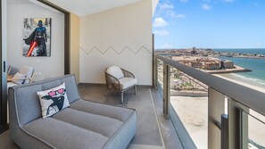 En balkong med utsikt över en strand och stadens silhuett, en grå soffa med en kudde och en fåtölj i rotting.