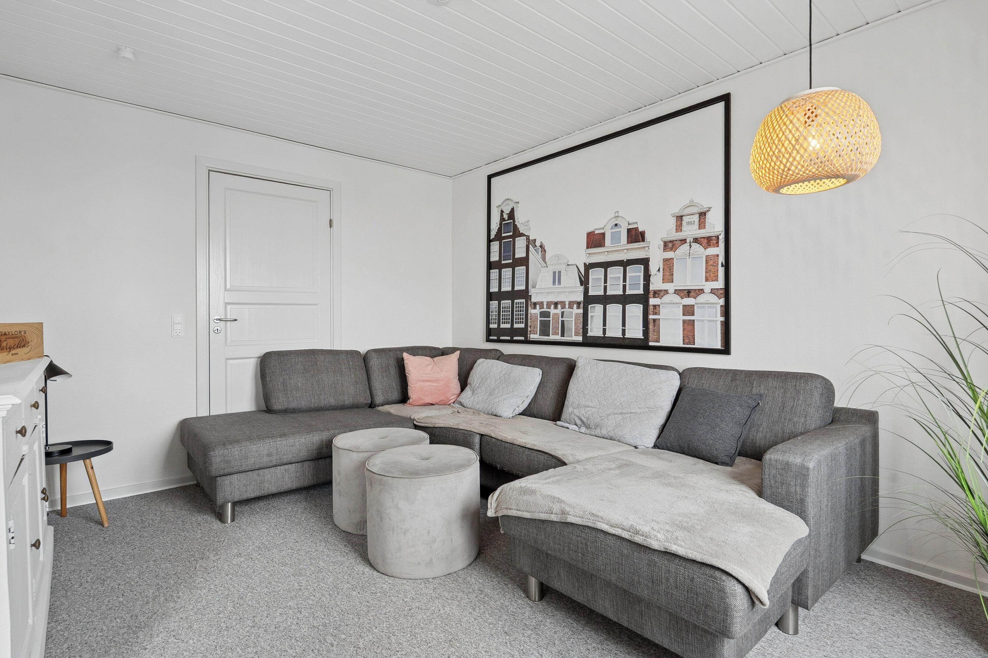 Un salon moderne avec un canapé d’angle gris, des poufs, une porte blanche et une œuvre d’art représentant un paysage urbain encadrée.