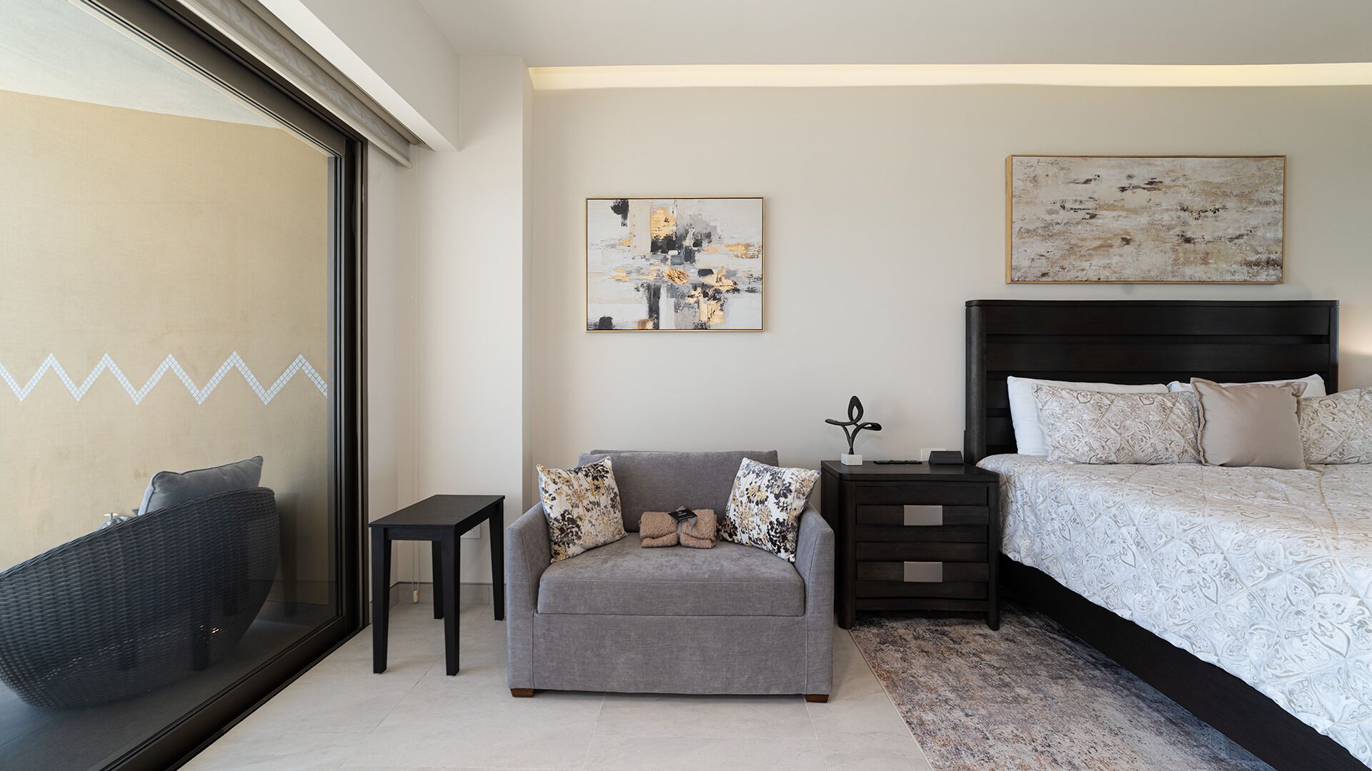 A modern bedroom with a large bed, a grey sofa, a small table, and artwork on the walls.