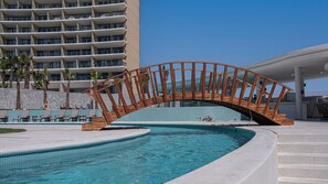 Ein modernes Hotel mit Poolbereich, das eine Holzbrücke, Liegestühle und ein großes Gebäude im Hintergrund zeigt.