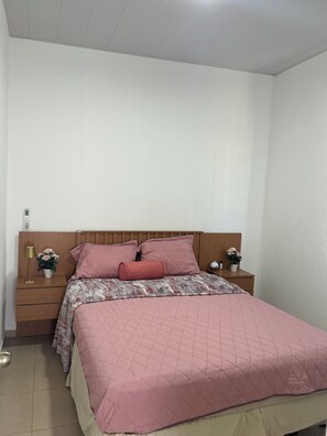 Une chambre à coucher avec une tête de lit en bois, un lit avec une literie rose, une table de chevet avec une lampe et un vase de fleurs, et une petite table de nuit avec une lampe et une bouteille.