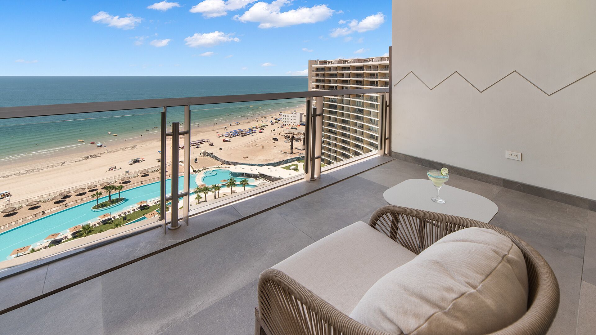 Ein Balkon mit Blick auf einen Strand und ein Schwimmbad, ein Korbstuhl und ein kleiner Tisch mit einem Getränk.