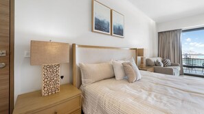 Ein Hotelzimmer mit einem großen Bett, einem Nachttisch mit Lampe, einem hölzernen Kopfteil und Meerblick durch das Fenster.
